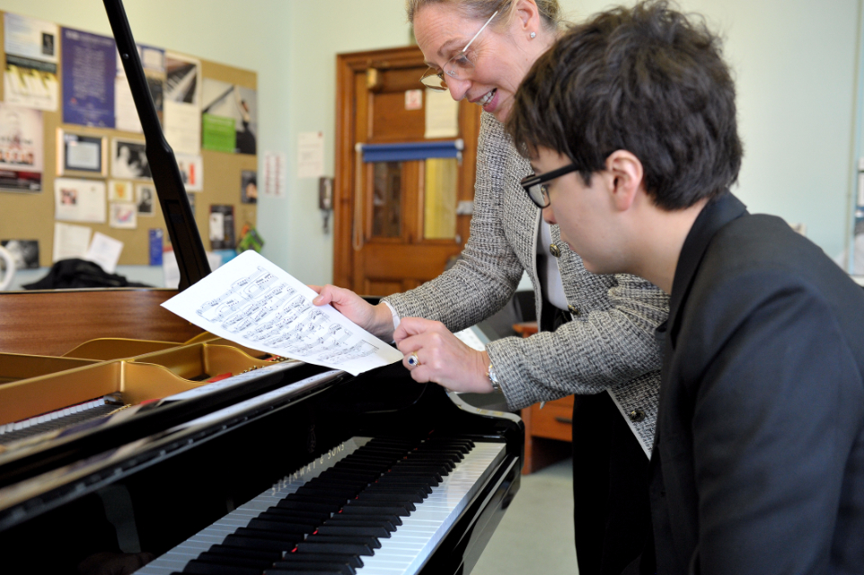 A male student looking at music sheets sitting at a piano, with a women with glasses talking to him, pointing at the music sheets.
